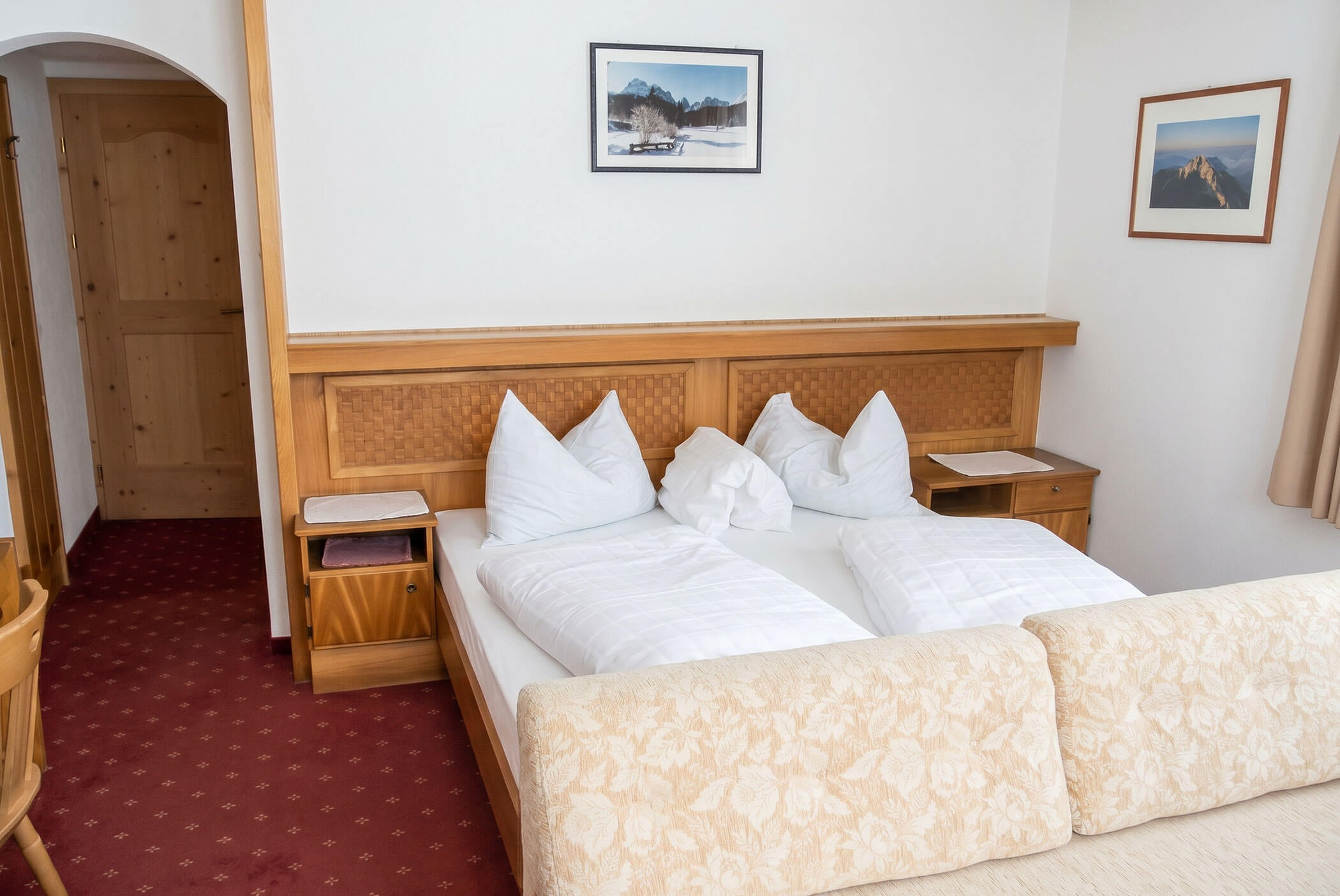 Ein ordentlich gemachtes Doppelbett mit weißem Bettzeug in einem gemütlichen Hotelzimmer mit Holzmöbeln und Kunstwerken an den Wänden. - Hotel Mooserhof