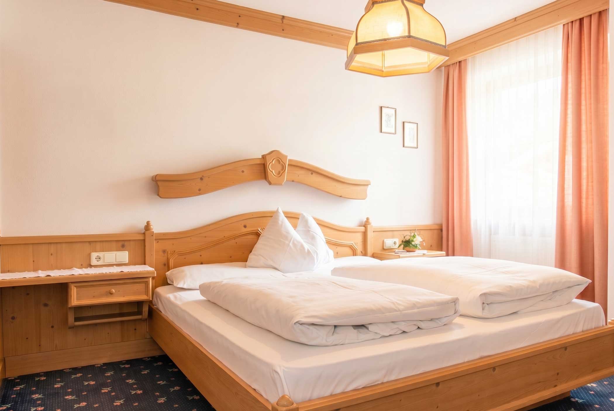 Gemütliches Hotelzimmer mit Holzmöbeln, einem Doppelbett, weißer Bettwäsche und Sonnenlicht durch transparente Vorhänge. - Hotel Mooserhof