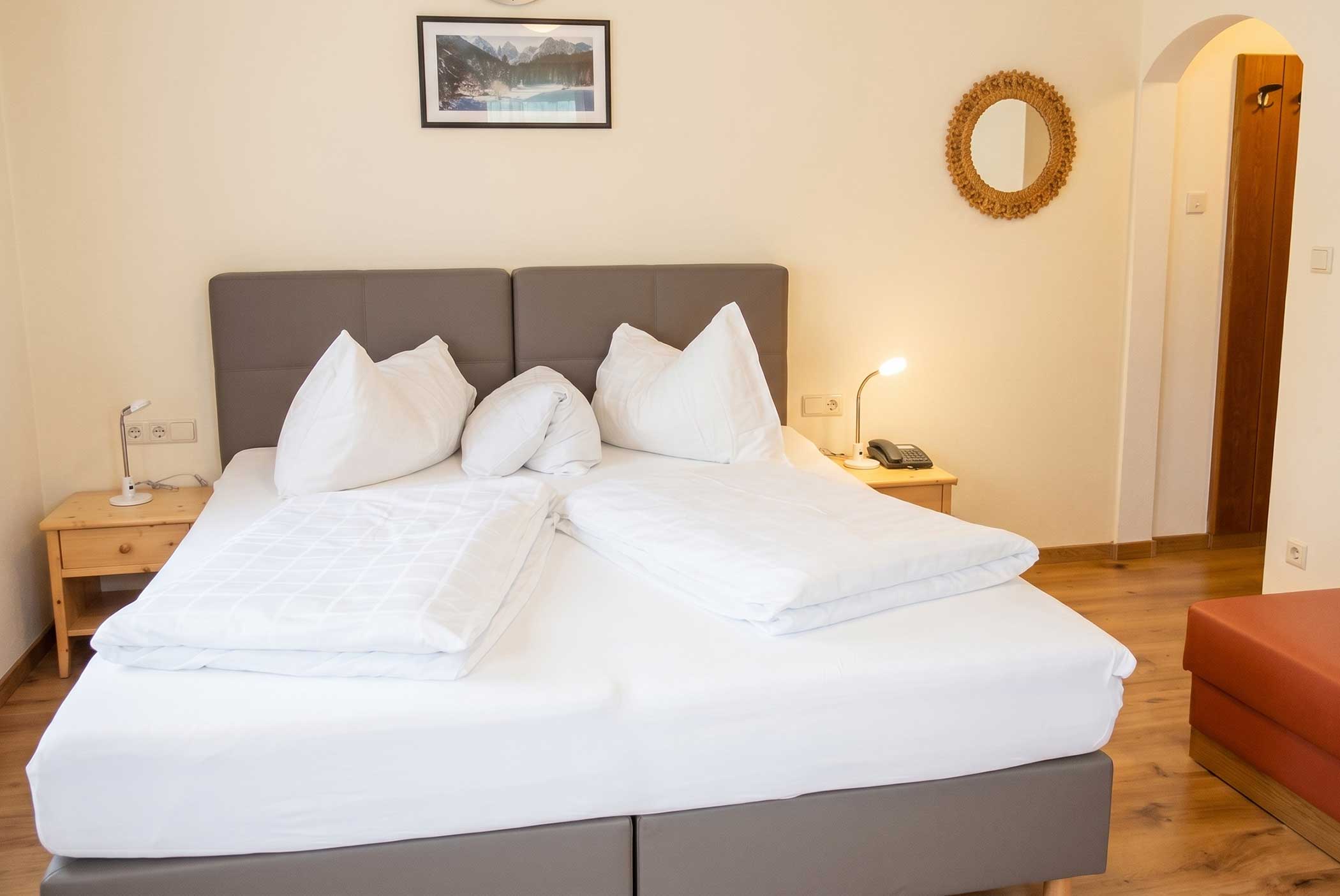 Schlafzimmer mit ordentlichem Doppelbett, Nachttischen mit Lampen und Wanddekoration über und neben dem Bett. - Hotel Mooserhof
