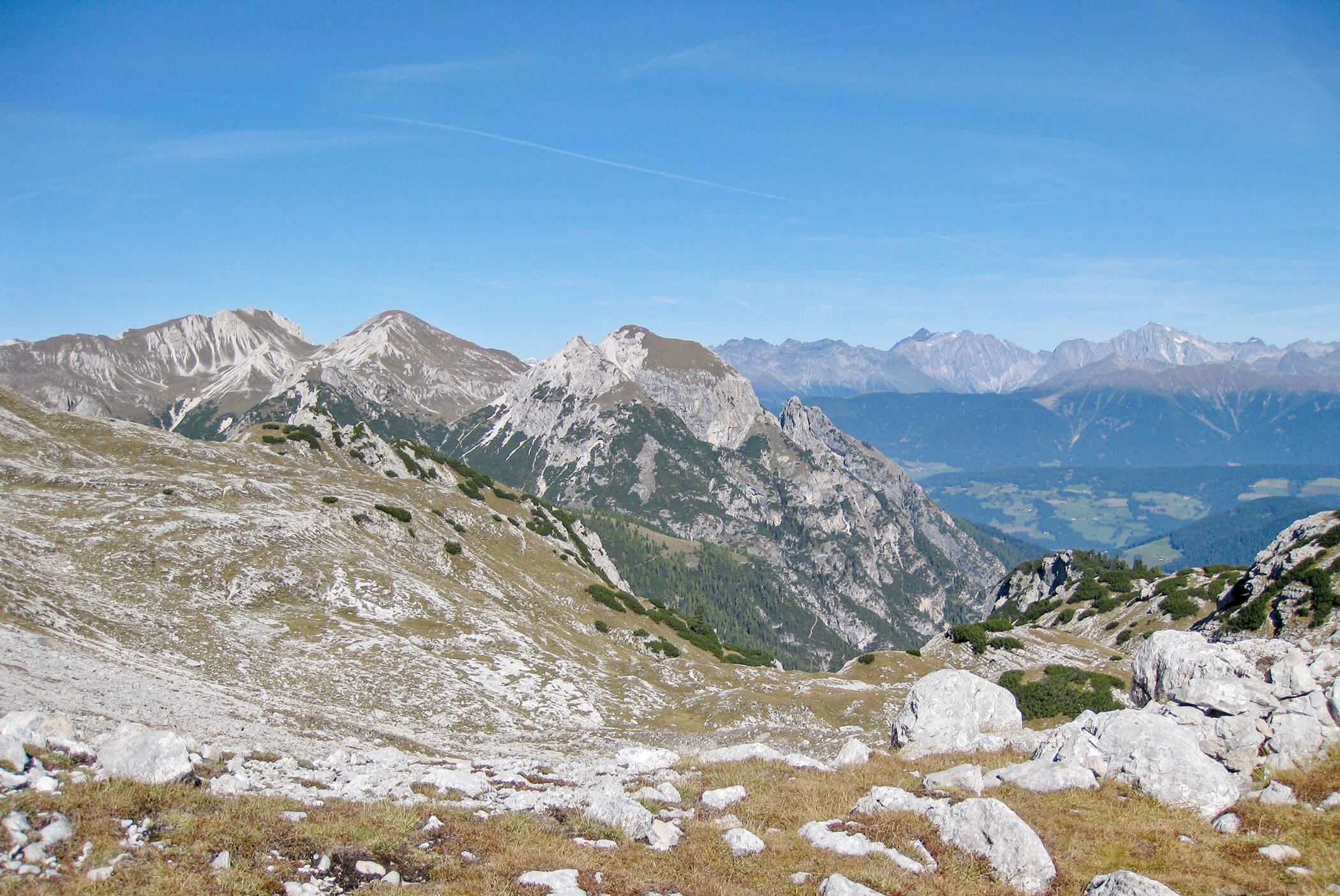 Felsige Gebirgslandschaft mit grasbewachsenen und felsigen Hängen unter einem klaren blauen Himmel, mit fernen Gipfeln im Hintergrund. - Hotel Mooserhof