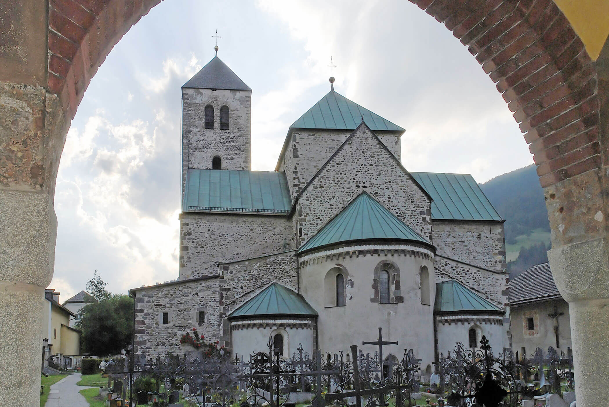 Steinkirche mit grünen Dächern durch einen Bogen gesehen, umgeben von Gräbern und Bergen im Hintergrund. - Hotel Mooserhof