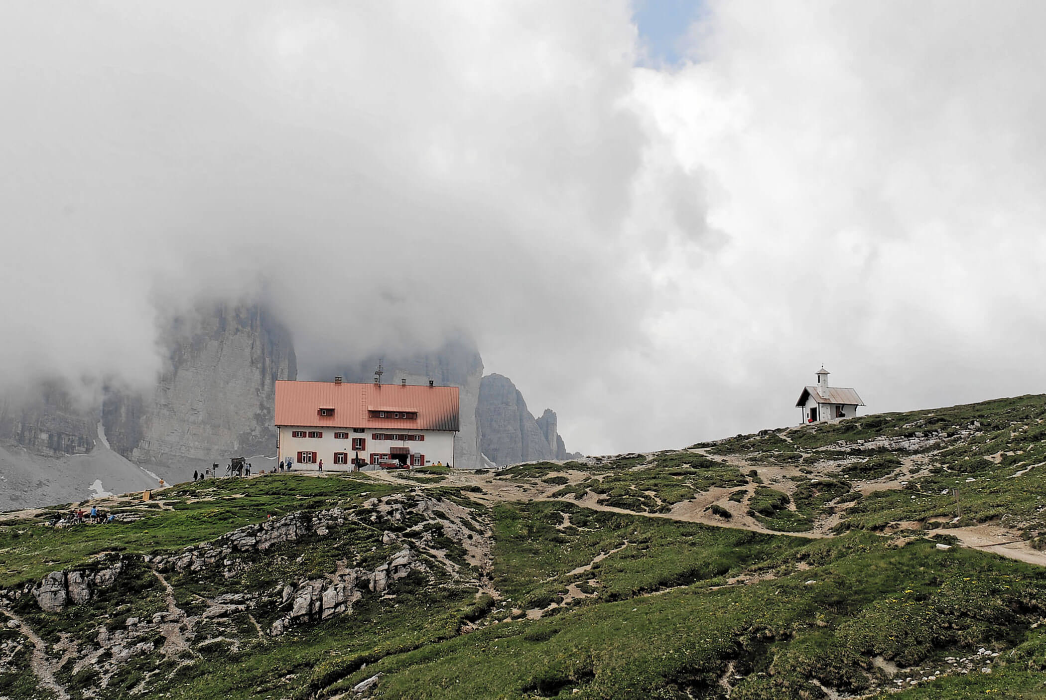 Eine Berghütte und eine kleine Kapelle liegen auf einem grasbewachsenen Hügel unter einem wolkenverhangenen Himmel, mit felsigen Gipfeln im Hintergrund. - Hotel Mooserhof