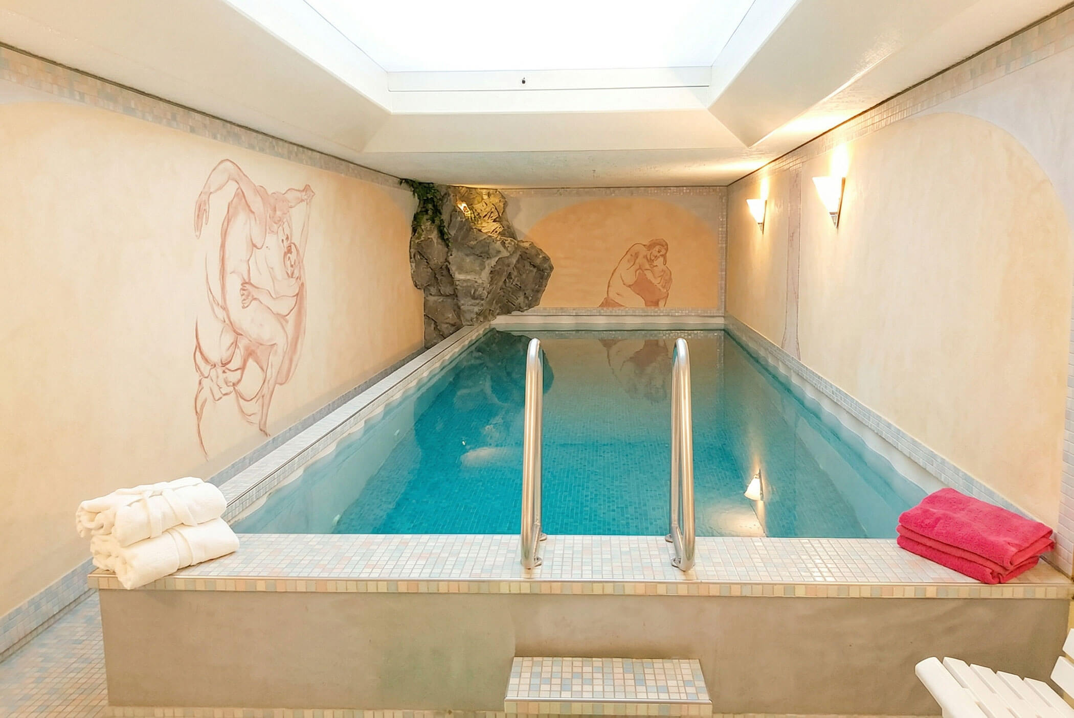 Hallenbad mit Handläufen, Oberlicht, Wandmalereien an den Wänden und gefalteten Handtüchern am Beckenrand. - Hotel Mooserhof